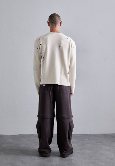 Maglione a trecce color crema con macchie nere, abbinato a pantaloni cargo scuri, larghi, con tasche laterali e orlo a polsino.