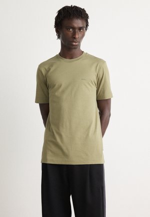 Man with short dreadlocks wearing an olive green BOSS t-shirt and loose black pants, standing with hands behind his back.