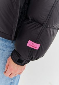 Czarna kurtka puchowa z różową metką z napisem „NALEŻY CI SIĘ NAJLEPSZE.” Zbliżenie na ramię ukazujące fakturę tkaniny i fason rękawa.