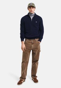 Granatowy sweter wykonany na drutach z zamkiem przy kołnierzu, szary golf pod spodem, beżowe spodnie z korduroy i brązowe trampki z pomarańczowymi akcentami. Wyeksponowane logo Caps.