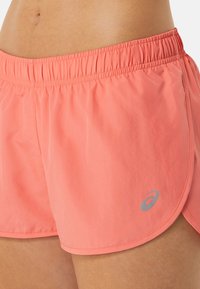 Primer plano de pantalones cortos deportivos de color coral con cinturilla elástica y pequeño logo gris de la marca en el lado izquierdo, usados por una persona.