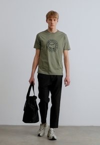 Olívazöld pamut póló kör alakú grafikával, fekete laza szabású nadrág és fekete táska. A modell fehér tornacipőt visel. Egyszerű, hétköznapi stílus.