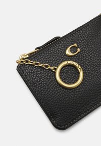 Portafoglio in pelle nera con finitura testurizzata, dotato di una catena in tonalità oro e hardware ad anello circolare, chiusura a zip nella parte superiore.