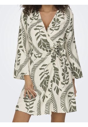 Femme portant une robe portefeuille mi-longue avec des motifs de feuilles vertes, des manches longues, et une ceinture nouée à la taille sur un tissu crème.