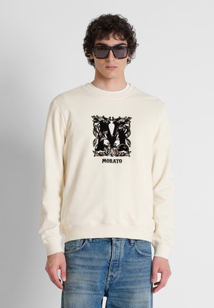 Joven con gafas de sol cuadradas negras, una sudadera color crema con decoración negra "M" y texto "Morato", y vaqueros azules.