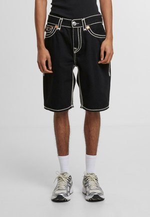 Shorts en denim noir avec des coutures blanches, cinq poches et une fermeture à bouton. Portés avec des chaussettes blanches et des baskets grises. Coupe régulière.