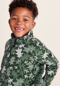 Fleece jacket with a green and gray leaf pattern, high collar, and zip front. Features a black logo patch on the sleeve.