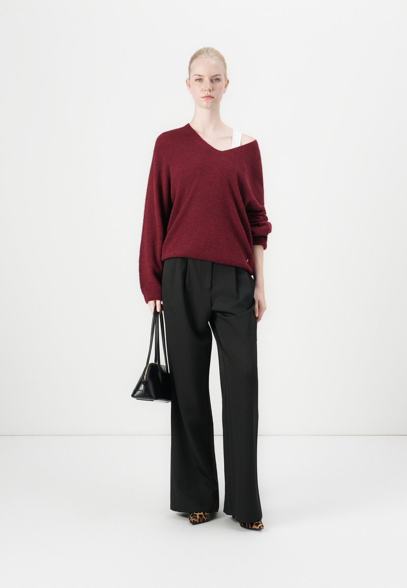 Burgunderfarbener, übergroßer Strickpullover mit V-Ausschnitt, kombiniert mit schwarzen weit geschnittenen Hosen. Accessoires sind eine kleine schwarze Tasche und Schuhe mit Leopardenmuster.