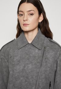 Veste en faux suède grise avec un grand col, fermeture éclair à l'avant et poches latérales, dotée de finitions métalliques et d'une texture douce.