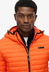 Chaqueta acolchada naranja con capucha y cierre de cremallera. Presenta detalles en negro y un logotipo de la marca en el pecho. Textura suave y diseño ajustado.