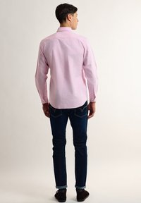 Camisa de botones rosa claro con mangas largas, yugo trasero y dobladillo redondeado, combinada con jeans azul oscuro y zapatos marrones.