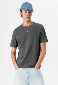 Graues Baumwoll-T-Shirt mit Rundhalsausschnitt und kurzen Ärmeln, mit einem kleinen bestickten Design auf der Brust. Kombiniert mit hellblauen Jeans und einer Jeansmütze.