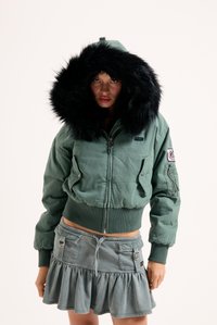 Zelená bomber bunda s čiernou kožušinou na kapucni, so zipsom a prednými vreckami; skombinovaná s rozšírenou denimovou sukňou vo svetlejšej farbe.