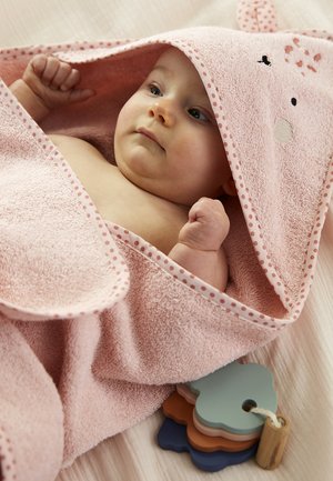 Baby wrapped in pink hooded towel with animal ears, lying on bed near colorful wooden toy shaped like stacked trees.