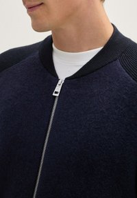 Ciemnoniebieska bomberka o gładkiej wełnianej fakturze, z prążkowanym kołnierzem i srebrnym zamkiem błyskawicznym. Posiada teksturowane akcenty na ramionach.