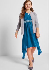 Abito teal con orlo alto-basso, vita decorata, abbinato a un cardigan grigio chiaro e a ballerine grigie, che mostra una texture di tessuto liscia.
