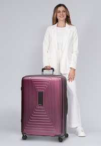 Hartschalenkoffer in glänzender burgunderroter Farbe mit einem strukturierten Wellenmuster, ausziehbarem Griff und vier Rädern für Mobilität.