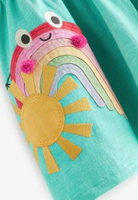 Tissu turquoise vif avec un soleil cousu et un arc-en-ciel souriant aux joues ornées de pompons, brodés sur la manche.