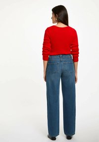 Maglione rosso leggero con maniche lunghe abbinato a jeans in denim blu a vita alta e gamba larga, con tasche posteriori e una texture liscia.