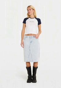 Weiße, kurz geschnittene T-Shirt mit marineblauen Raglanärmeln, das ein einfaches Grafikdesign zeigt, kombiniert mit einem hellblauen Jeans-Bleistiftrock und schwarzen kniehohen Stiefeln.