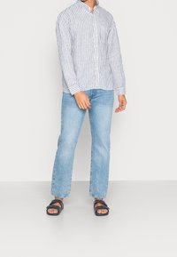 Camicia a righe blu e bianca, jeans dritti azzurri chiari e sandali neri. Vestibilità casual con colletto a punta e dettagli sulla tasca.