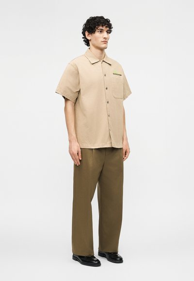 Άνδρας που στέκεται με μπεζ κοντομάνικο πουκάμισο με κουμπιά και χακί παντελόνι φαρδιάς γραμμής, φορώντας μαύρα παπούτσια, μπροστά σε απλό φωτεινό φόντο.