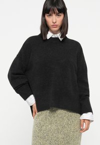 Svart oversized genser med myk tekstur, ribbede mansjetter og rund hals, lagd over en hvit skjorte med krage og et mønstrete skjørt.