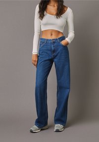 Jeans in denim blu con una vestibilità a vita alta e gambe larghe. Indossati con un top bianco a maniche lunghe e a coste, corto, e sneakers grigie.