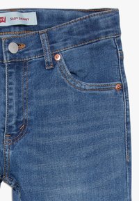 Detailní záběr modrých džínoviny ukazující přední kapsu, měděné nýty, oranžové prošívání, knoflík a štítek "510™ SKINNY".