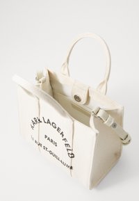 Mała beżowa torebka z płótna z krótkimi uchwytami, czarny napis logo "Karl Lagerfeld Paris" z przodu oraz zapięcie na zatrzask, lekko otwarta.