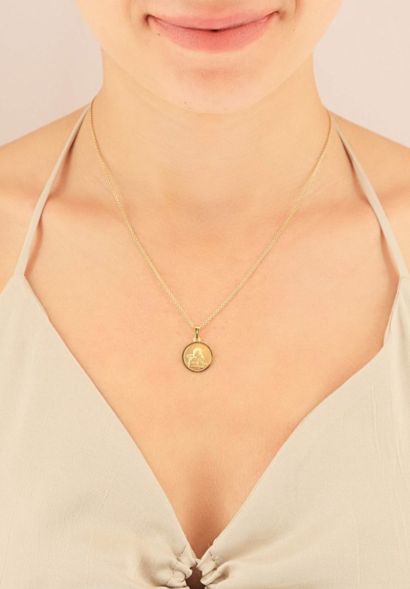 Collier en or avec un pendentif rond gravé en détail, suspendu à une chaîne délicate, porté sur un haut en tissu léger.