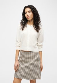 Γυναίκα με σγουρά μαλλιά, φορώντας λευκό ζακέτα με κουμπιά και μπεζ καρό φούστα, στέκεται μπροστά σε απλό φωτεινό φόντο.