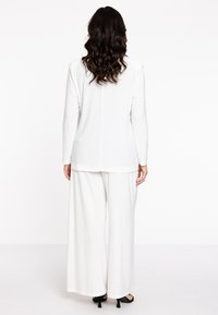 Un haut blanc à manches longues assorti à un pantalon large, tous deux en tissu doux. Détail notable : fente au dos du haut, porté avec des talons noirs.