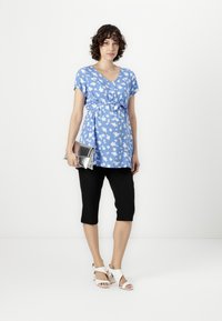 Tunic floreale blu con scollo a V, vita cintrata, abbinata a pantaloni capri neri e tacchi bianchi con cinturini. Borsetta argentata in mano.