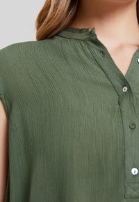 Blusa verde oliva con un tejido texturizado y arrugado. Presenta un escote abotonado y un cuello con forma de onda. Mangas cortas y botones sutiles.