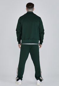 Zelený športový teplákový súprava z jemnej textúrovanej látky. Bunda má vysoký golier; nohavice majú bočné akcenty s kvetinovými vzormi.