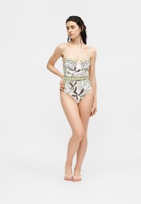 Femme aux cheveux foncés mouillés portant un maillot de bain une pièce blanc à motifs floraux avec des accents verts et des sandales blanches, debout devant un fond uni.