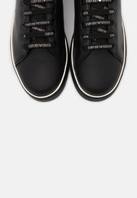 Černé kožené tenisky s bílým lemem podrážky a černými tkaničkami, na kterých se opakuje branding "Emporio Armani".