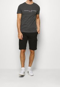 Mężczyzna w ciemnoszarej koszulce Tommy Hilfiger, czarnych szortach cargo, białych skarpetkach i białych sneakersach stoi na jednolitym tle.