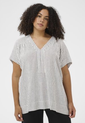 Kvinde med krøllet hår iført en hvid kortærmet V-hals top med tynde sorte lodrette striber og sorte bukser foran en ensfarvet baggrund.