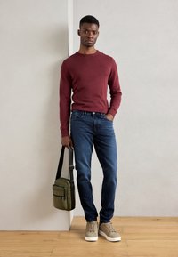 Maroon knit sweater, blue jeans, beige sneakers; holding an olive green bag with a shoulder strap. Smooth textures, casual fit.