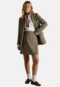 Súprava z karovaného materiálu v hnedých odtieňoch pozostávajúca z blejzera a sukne, doplnená bielou košeľou a vzorovaným modrým šálom. Hnedé mokasíny a biele ponožky do členkov dotvárajú celkový vzhľad.