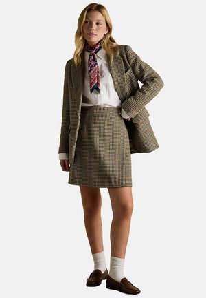 Karierte Blazer- und Rockkombination in Brauntönen, kombiniert mit einem weißen Hemd und einem gemusterten blauen Schal. Braune Mokassins und weiße Kniestrümpfe vervollständigen den Look.