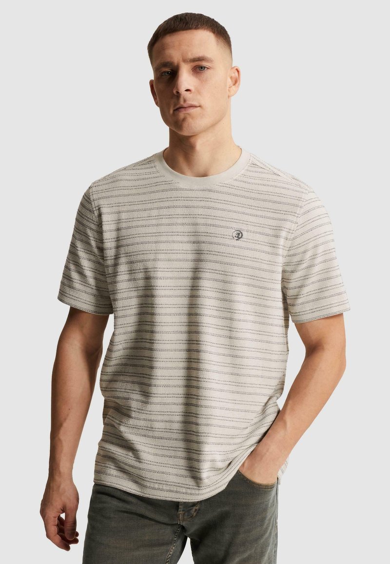 Jeune homme portant un t-shirt rayé gris clair à manches courtes avec un petit logo sur la poitrine, debout, une main dans la poche.