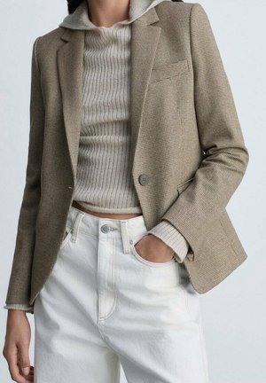 Blazer à carreaux beige sur un pull crème côtelé, associé à un jean blanc taille haute, une main dans la poche, style décontracté.