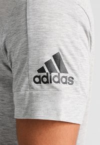 T-shirt grigia a maniche corte realizzata in tessuto misto cotone con una texture liscia, caratterizzata da un logo Adidas nero sul retro del manico sinistro.