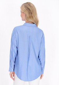 Camicia blu chiaro con bottoni, a righe verticali bianche e arancioni, maniche lunghe e una piega sul retro per una maggiore libertà di movimento. Tessuto di cotone liscio.
