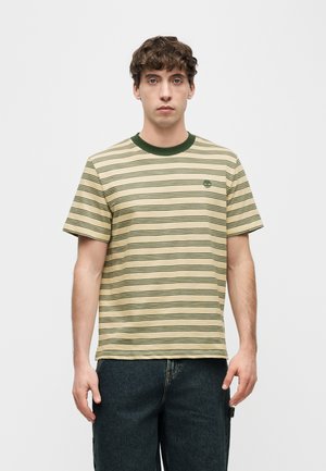 Ung mann med kort krøllete hår iført beige og grønstripet kortermet T-skjorte og mørke jeans, stående mot en enkel lys bakgrunn.