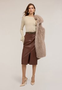 Beige Langarmshirt mit ausgestellten Ärmeln, brauner Leder-Bleistiftrock mit vorderem Schlitz, leichter Pelzmantel und hautfarbene High Heels.