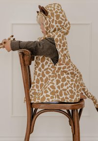 Kapucē ar žirafes rakstu, veidots no bēša un brūna auduma, ar ausīm un asti, ko valkā bērns, sēžot uz koka krēsla.